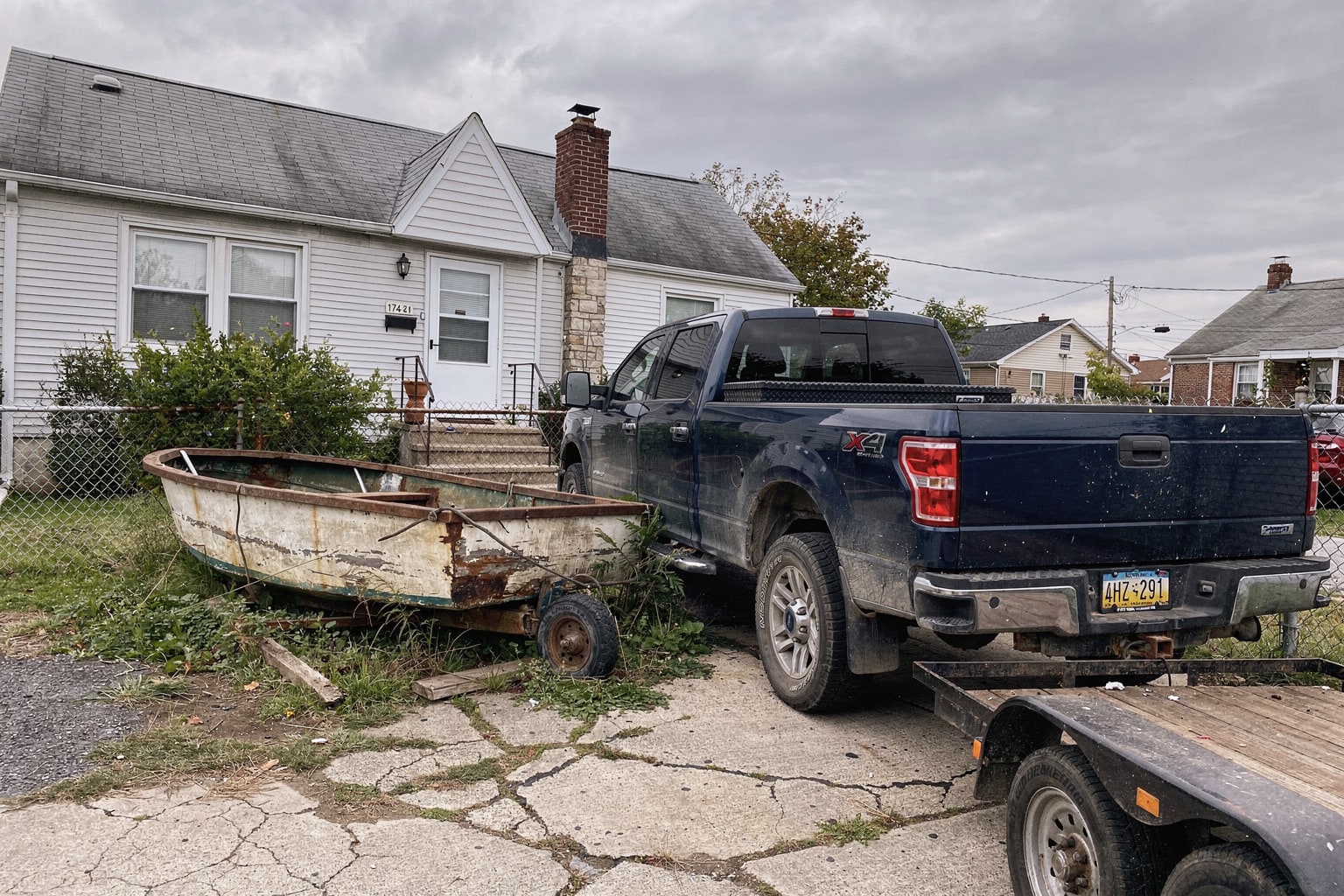 Abandoned junk boat on cracked concrete surrounded by weeds in Queens NY, pickup truck and trailer ready for disposal