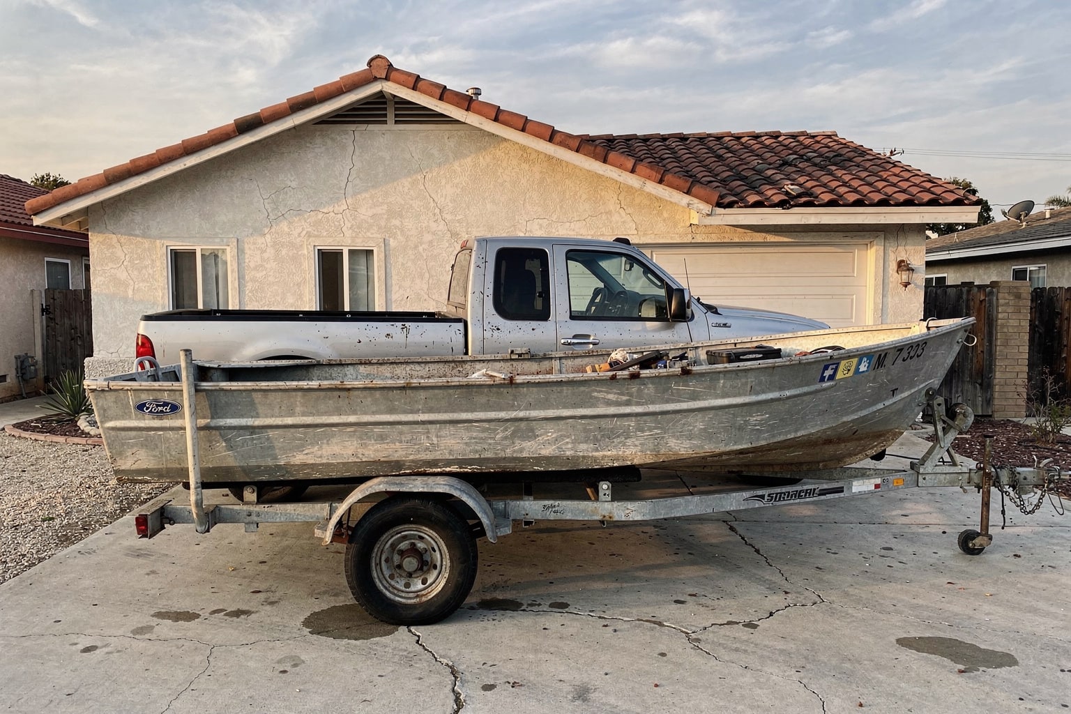 Weathered aluminum fishing boat on trailer secured behind pickup truck in California residential driveway, any condition salvage