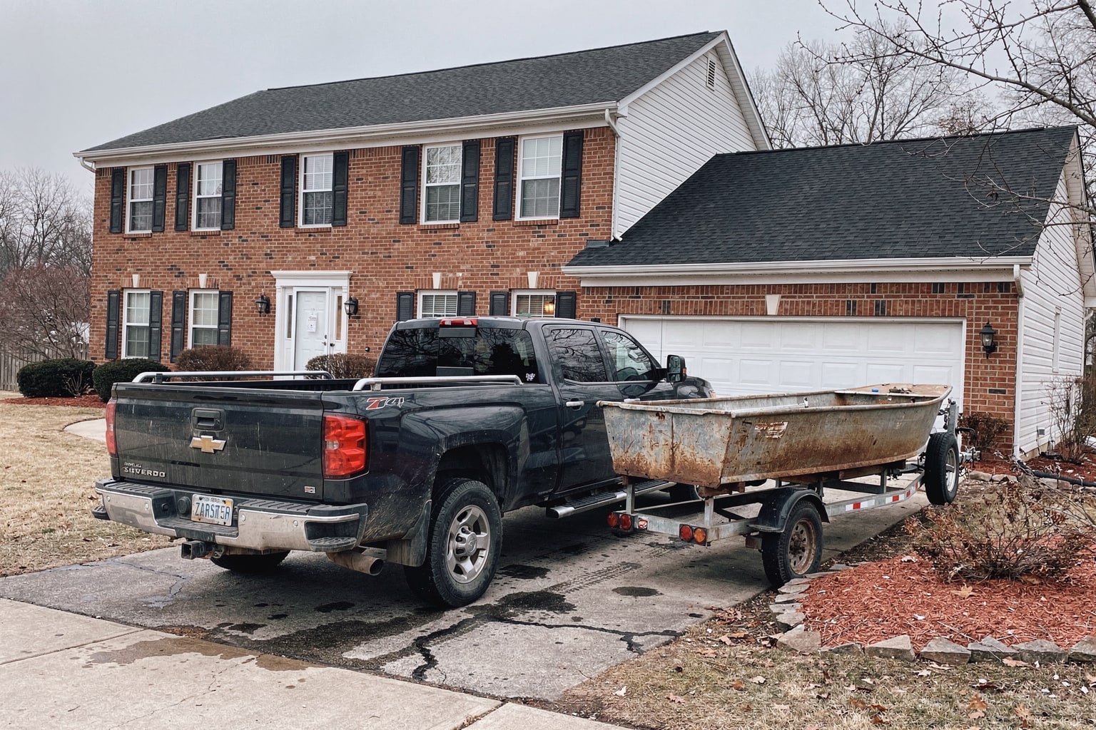 Junk boat removal Columbus Ohio on residential driveway with pickup truck and trailer, weathered aluminum fishing boat