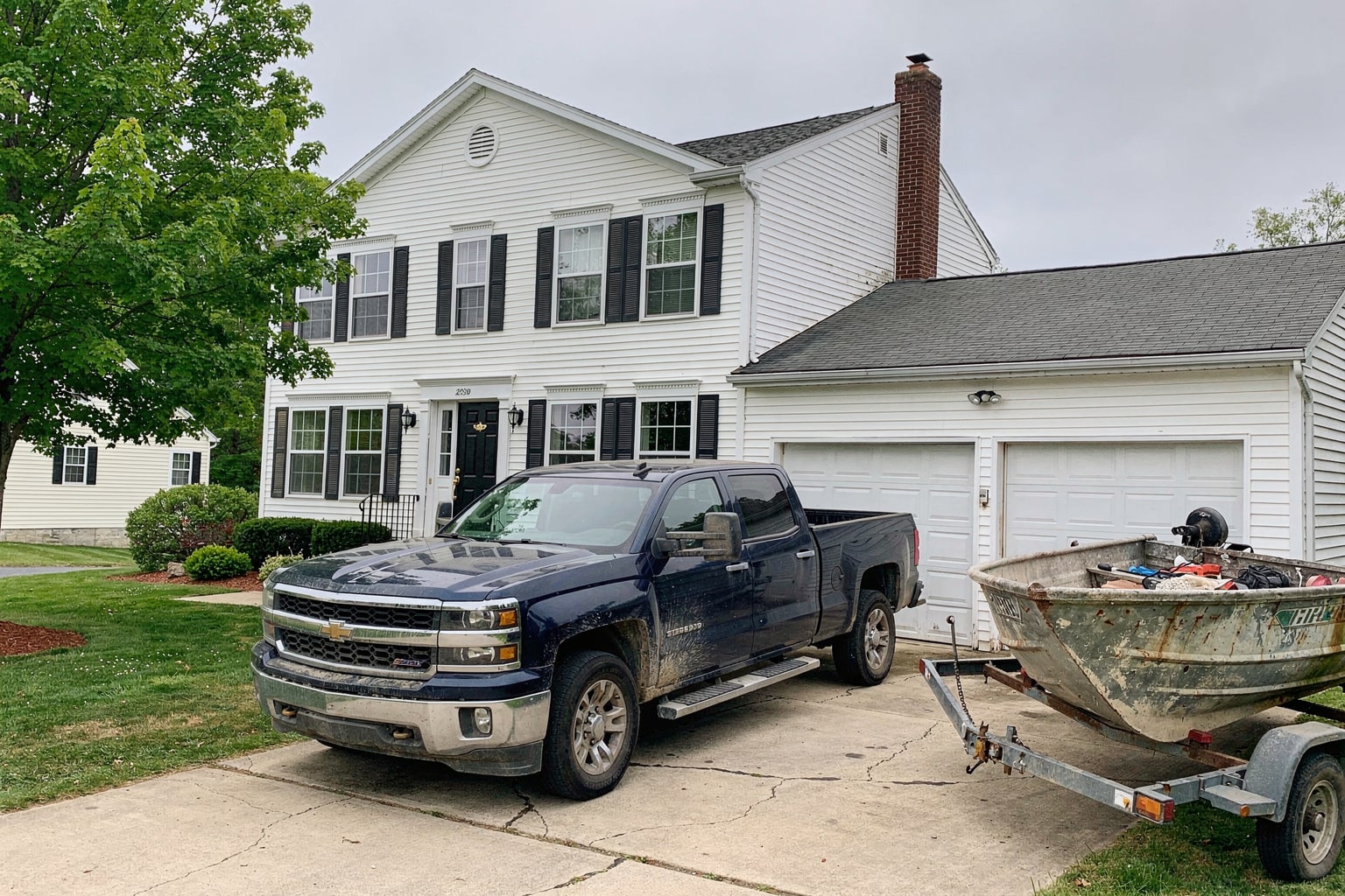 Dark blue pickup truck and boat trailer carrying weathered aluminum fishing boat on Connecticut residential driveway, colonial home background