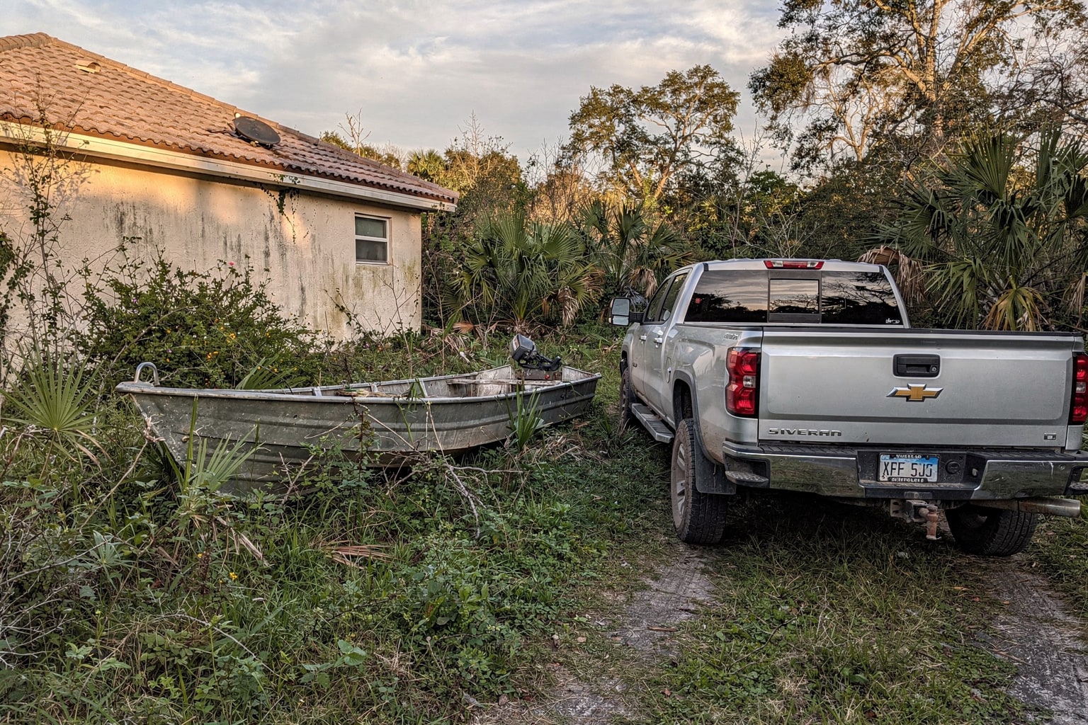 Abandoned 16-foot aluminum jon boat in overgrown Florida property surrounded by weeds and kudzu, pickup truck with boat removal trailer approaching for salvage