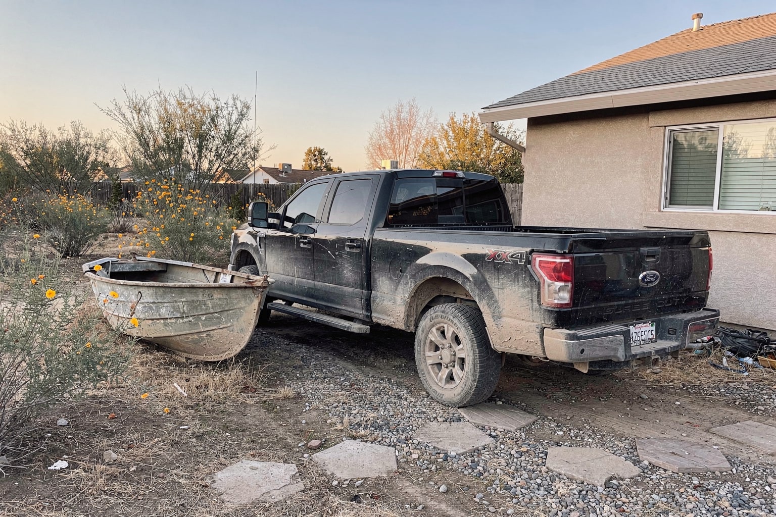 Abandoned junk boat removal Fresno, weathered jon boat in overgrown yard with pickup truck and empty trailer approaching for disposal