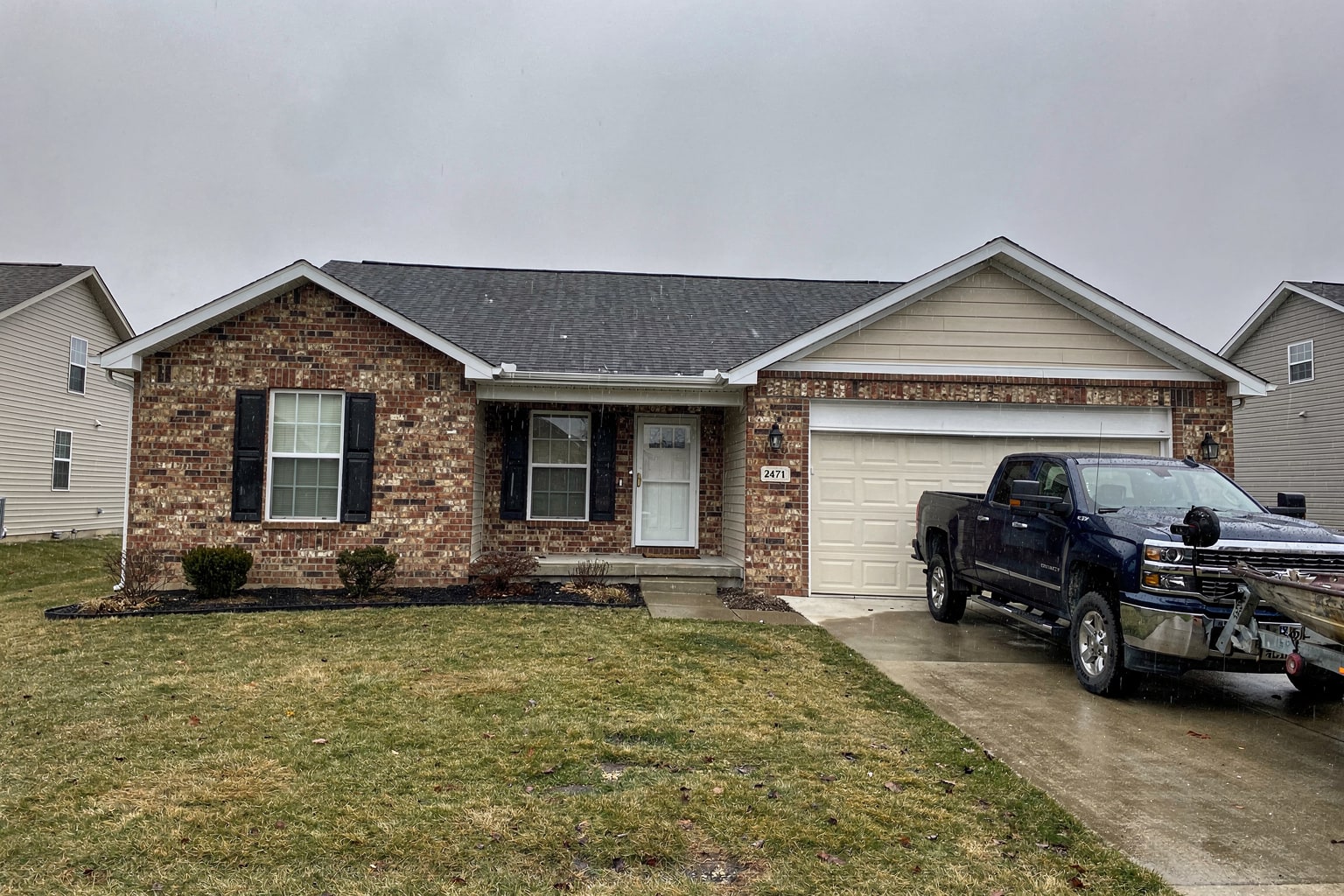 Dark blue pickup truck and boat trailer carrying weathered powerboat in Indiana residential driveway, brick home and mailbox visible
