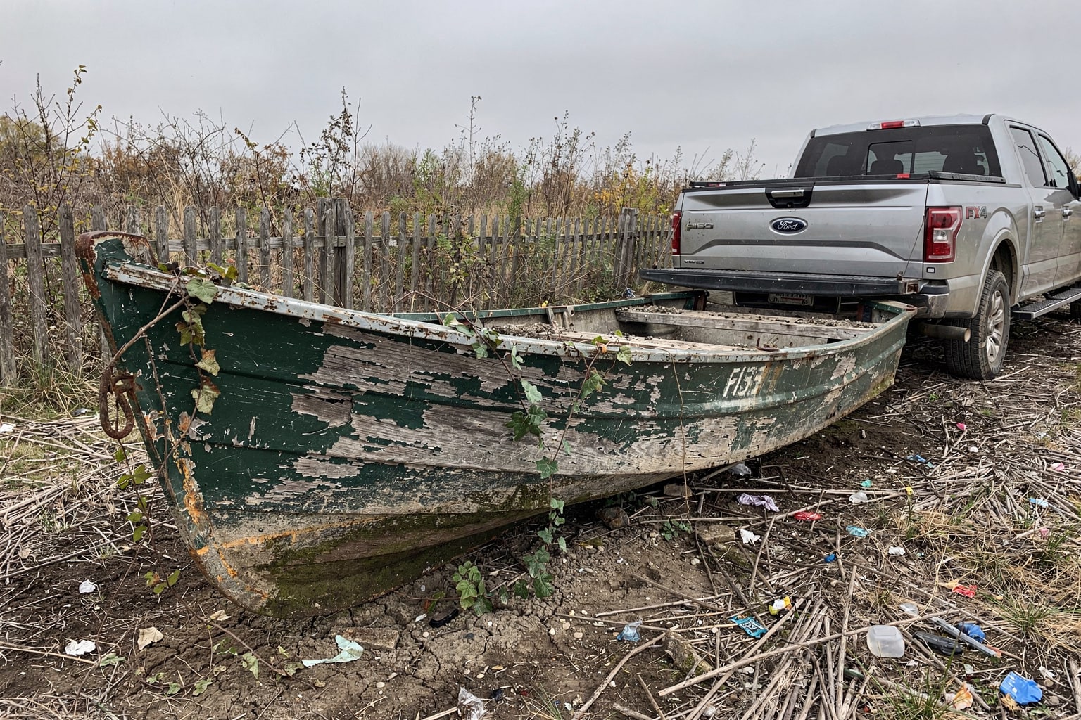 Rotted wooden jon boat on abandoned Iowa property surrounded by weeds, ready for marine salvage removal