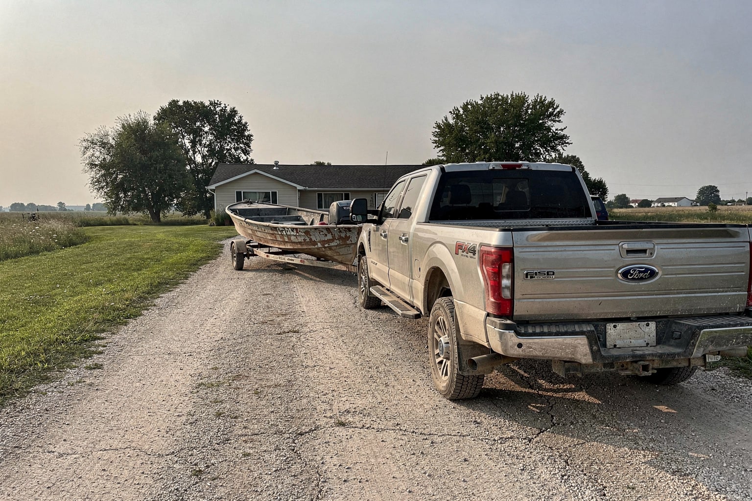 Weathered aluminum fishing boat on trailer attached to silver pickup truck in rural Iowa driveway for junk boat removal