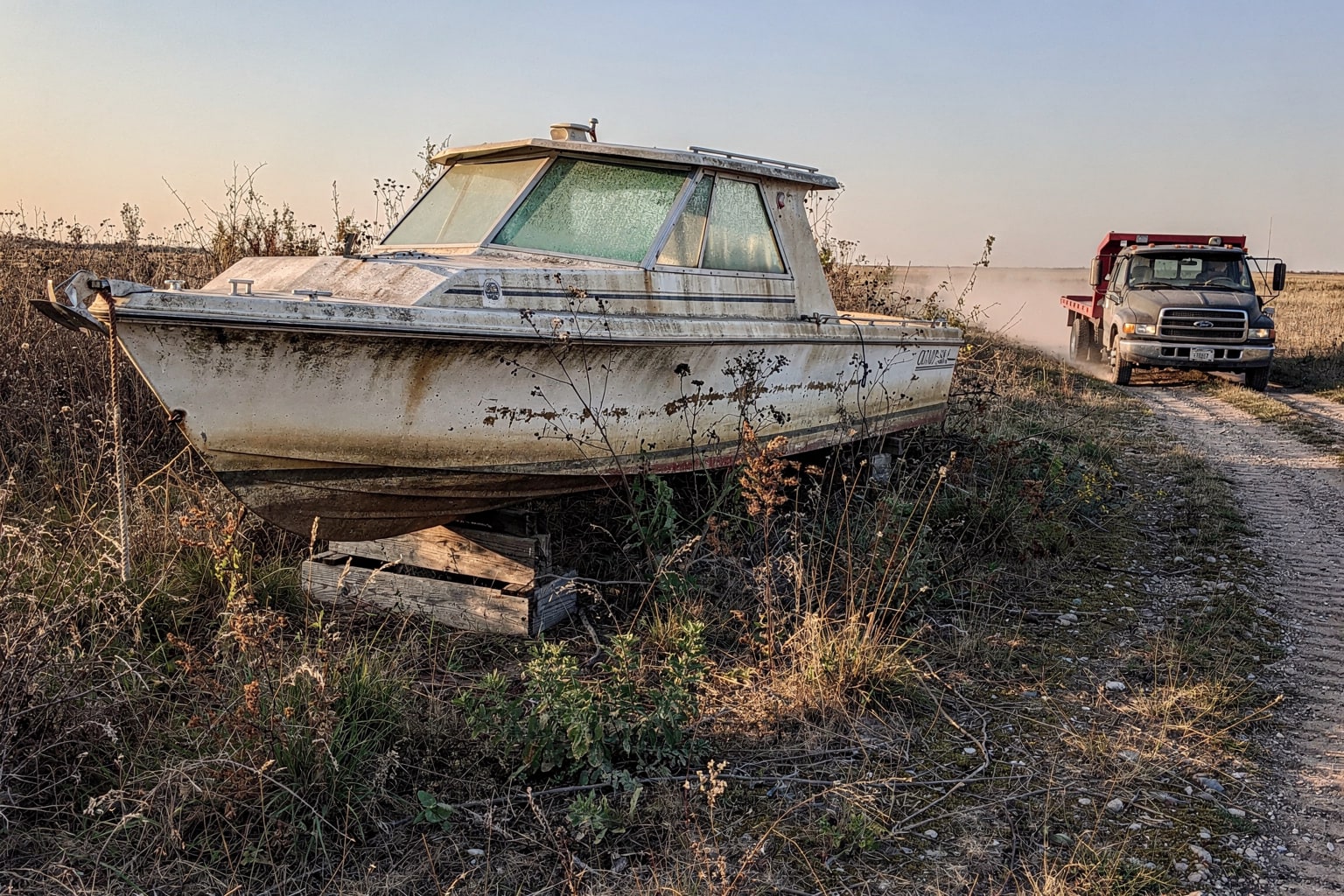 Abandoned deteriorated cabin cruiser on ground surrounded by overgrown prairie weeds, salvage truck approaching for Kansas junk boat removal