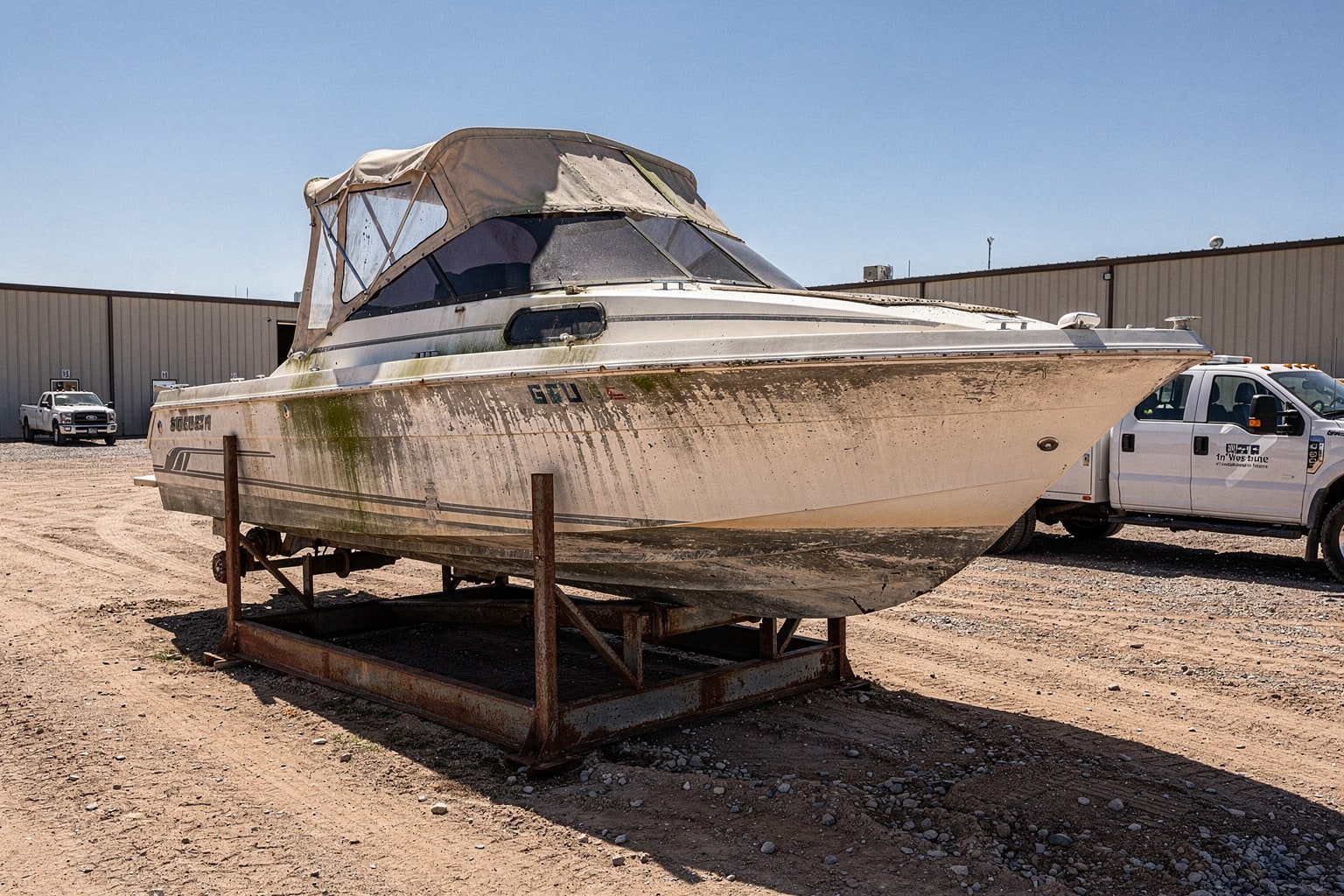 Large weathered cabin cruiser powerboat on steel cradle in Kansas salvage yard with industrial warehouse, professional marine disposal service