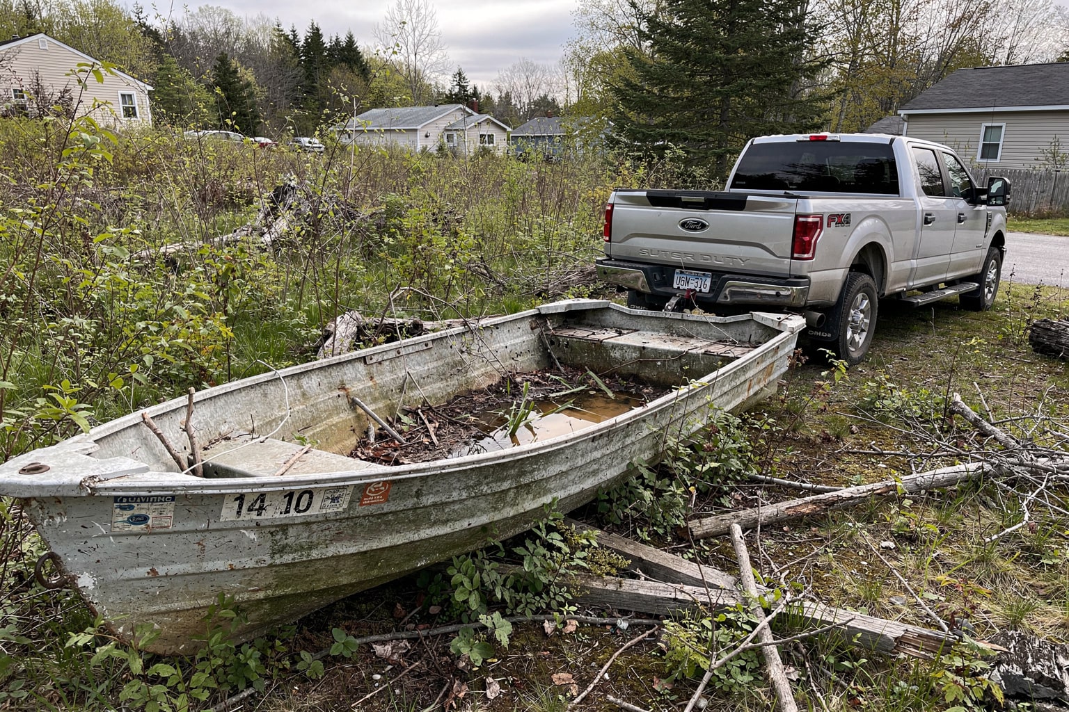 Abandoned jon boat in overgrown Maine backyard with pickup truck and trailer ready for junk boat removal