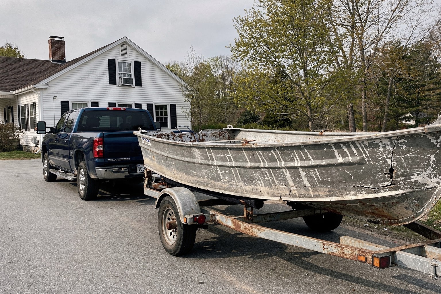 Severely damaged aluminum fishing boat loaded on trailer in Maine driveway with blue Chevy pickup truck