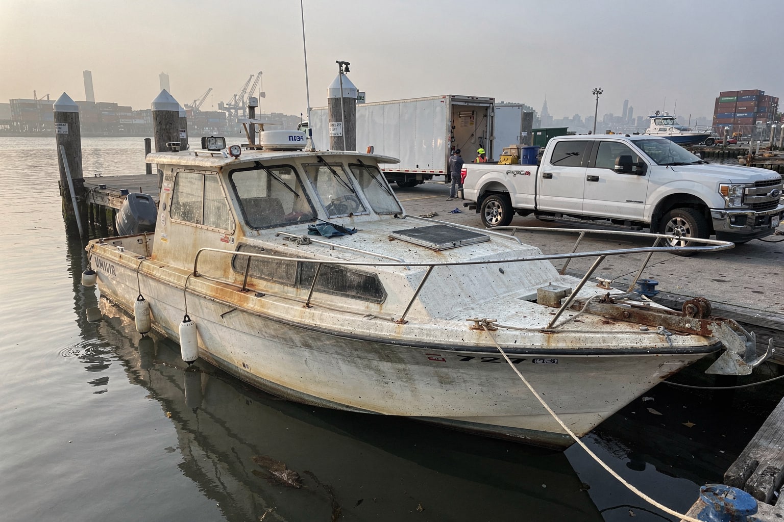 Dark blue pickup truck with boat removal trailer carrying worn fiberglass powerboat at Manhattan commercial dock