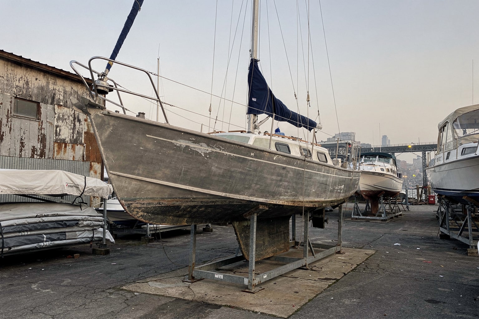 Weathered 28-foot aluminum sailboat on steel cradle at Manhattan boatyard with other stored boats visible