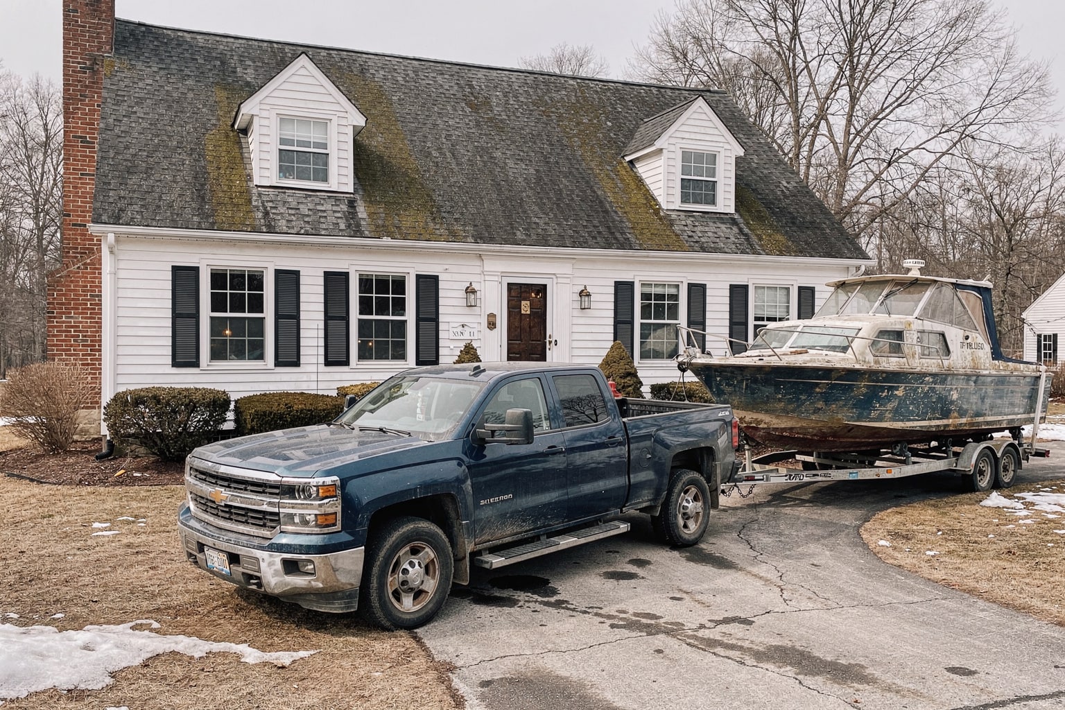 Large weathered cabin cruiser loaded on tandem-axle trailer behind blue Chevy pickup in Massachusetts residential driveway