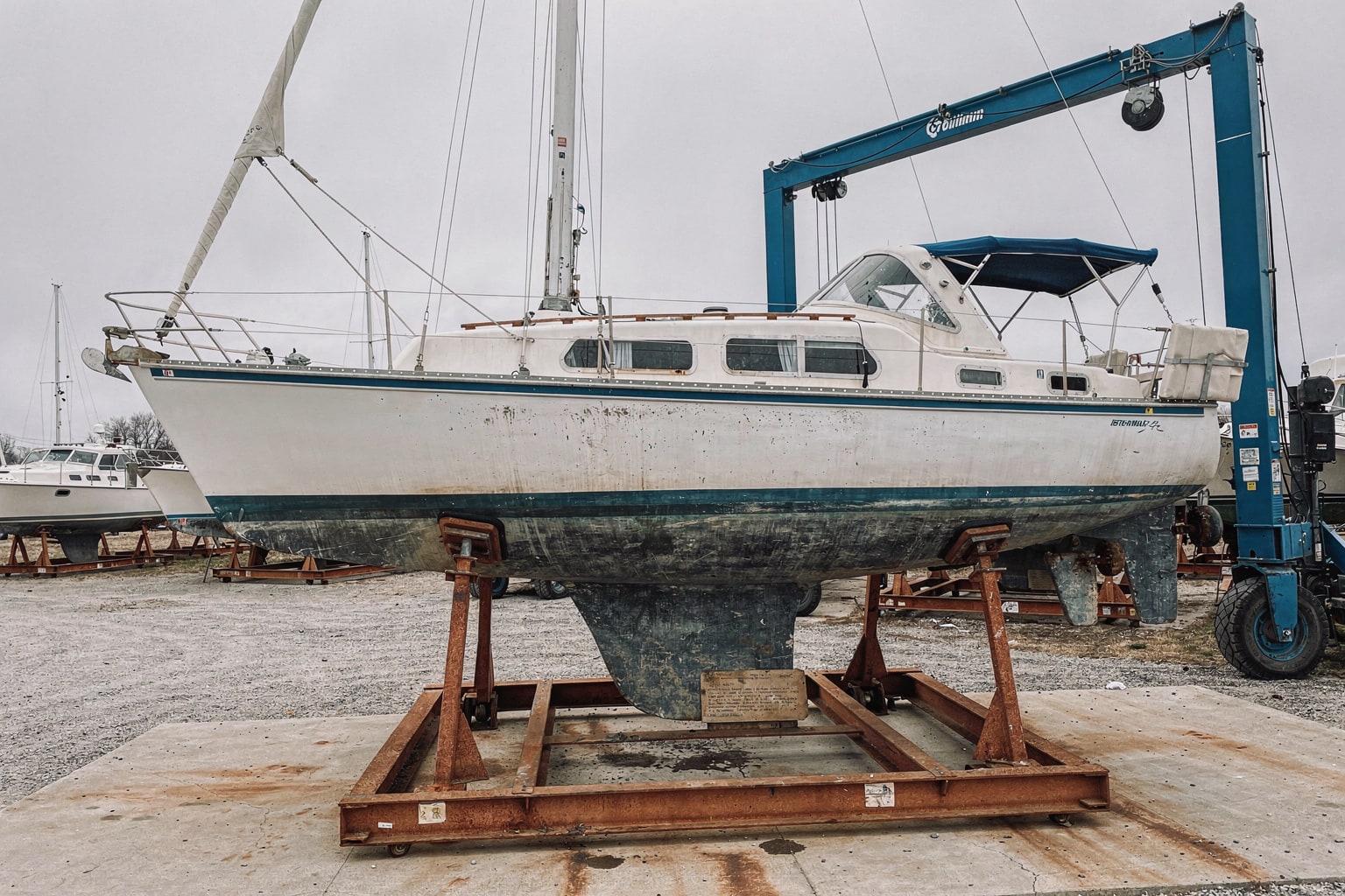 Large sailboat on steel cradle at Michigan boatyard with blue travel-lift gantry crane ready for marine salvage removal