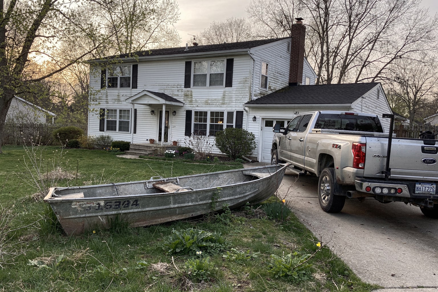 Abandoned junk boat removal Milwaukee backyard overgrown with weeds, silver truck with empty trailer ready for boat disposal