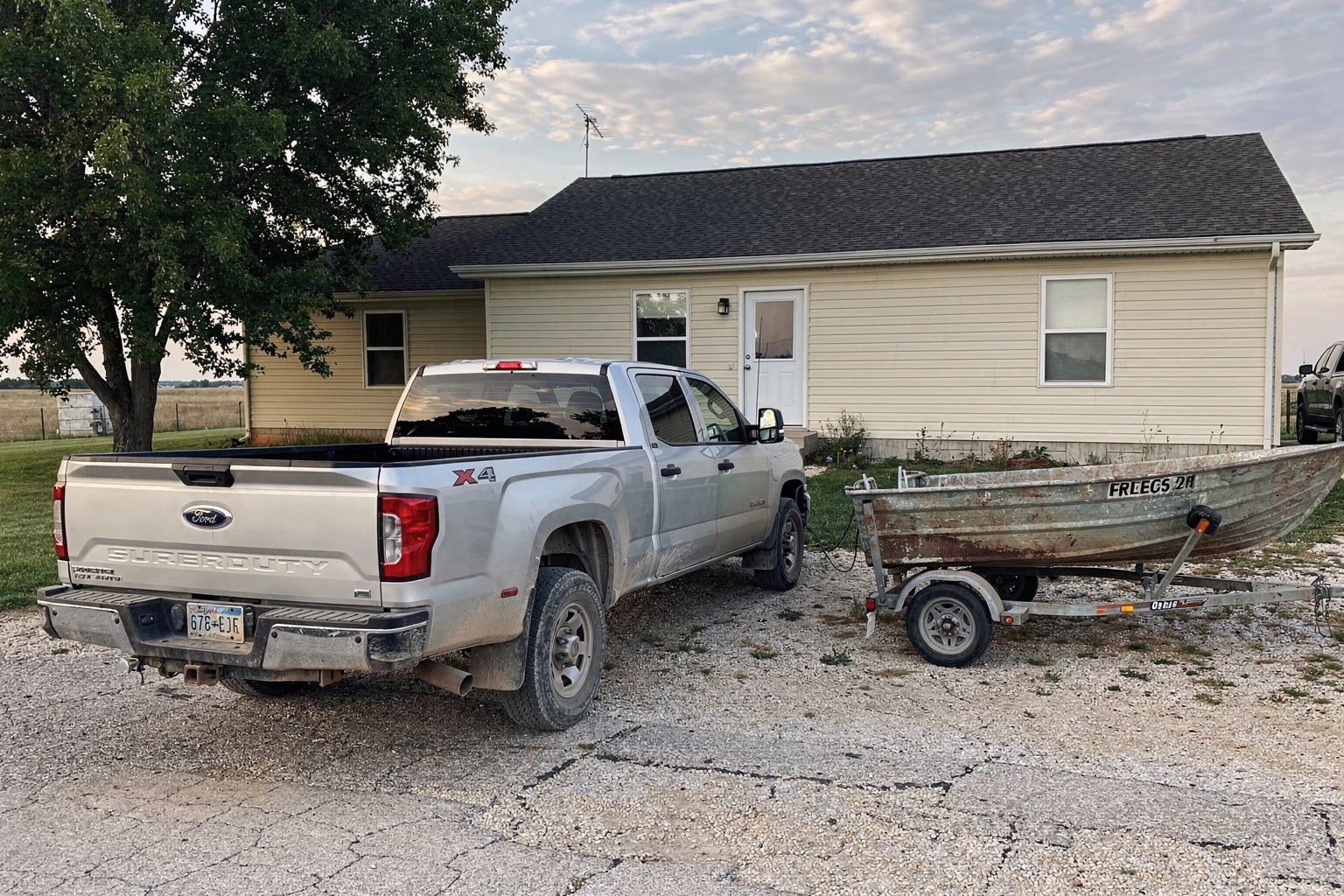Weathered aluminum fishing boat loaded on trailer behind silver pickup truck on Nebraska gravel driveway, ready for junk boat removal