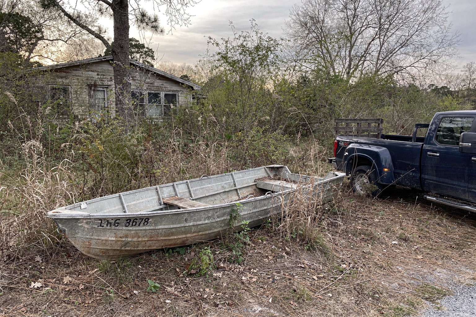 Abandoned 16-foot aluminum jon boat in overgrown North Carolina lot with pickup truck and empty trailer ready for salvage removal