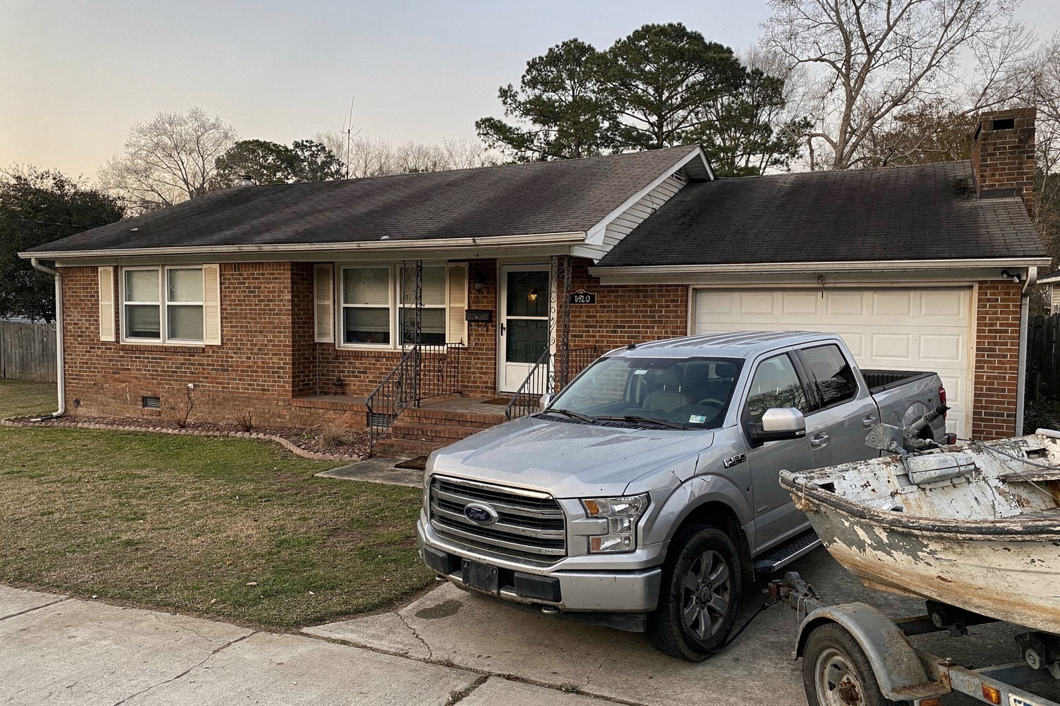Weathered 22-foot powerboat loaded on tandem-axle trailer behind dark blue pickup truck on North Carolina residential driveway near house