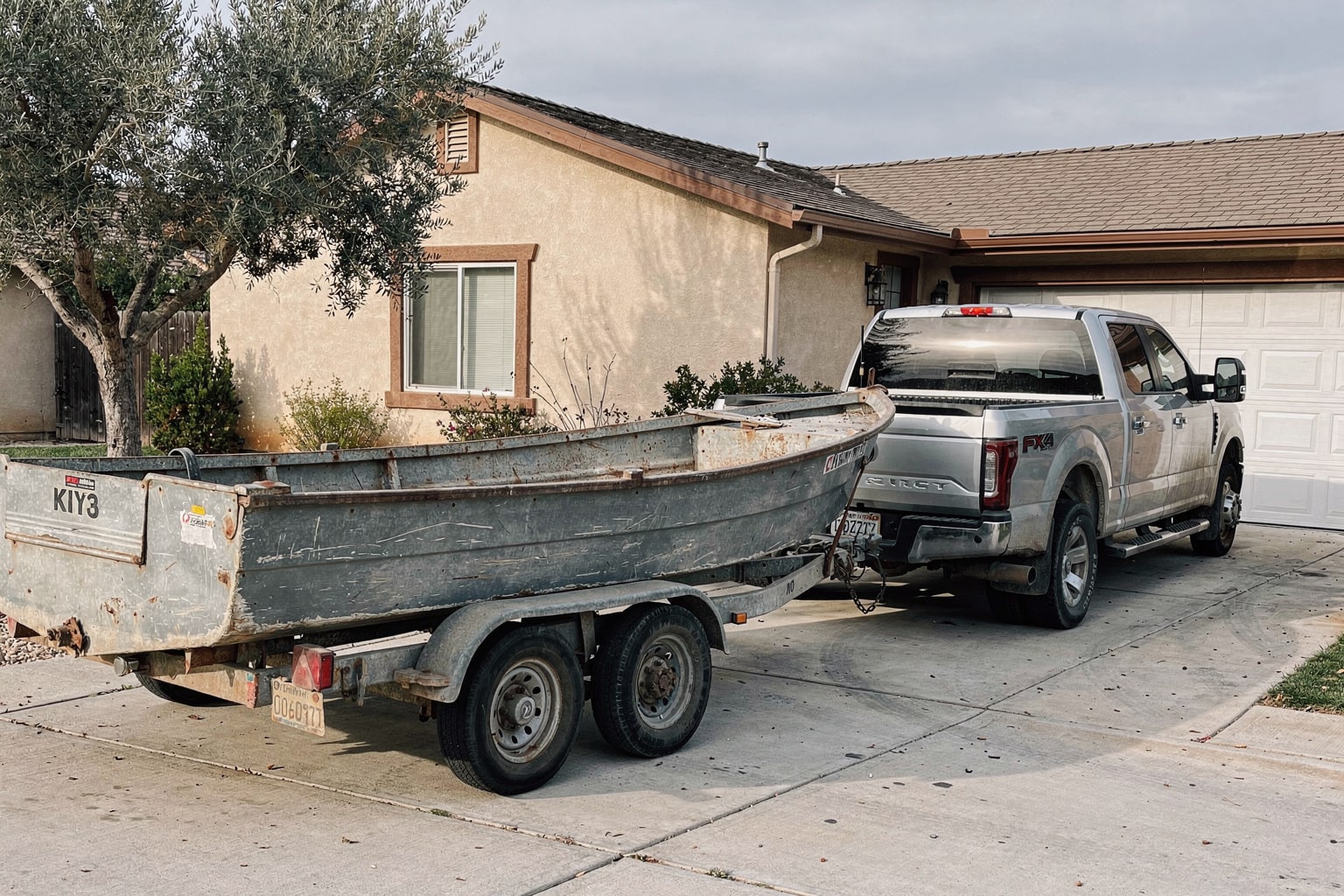 Junk boat removal Oakland CA — weathered aluminum boat loaded on trailer behind pickup truck in residential driveway