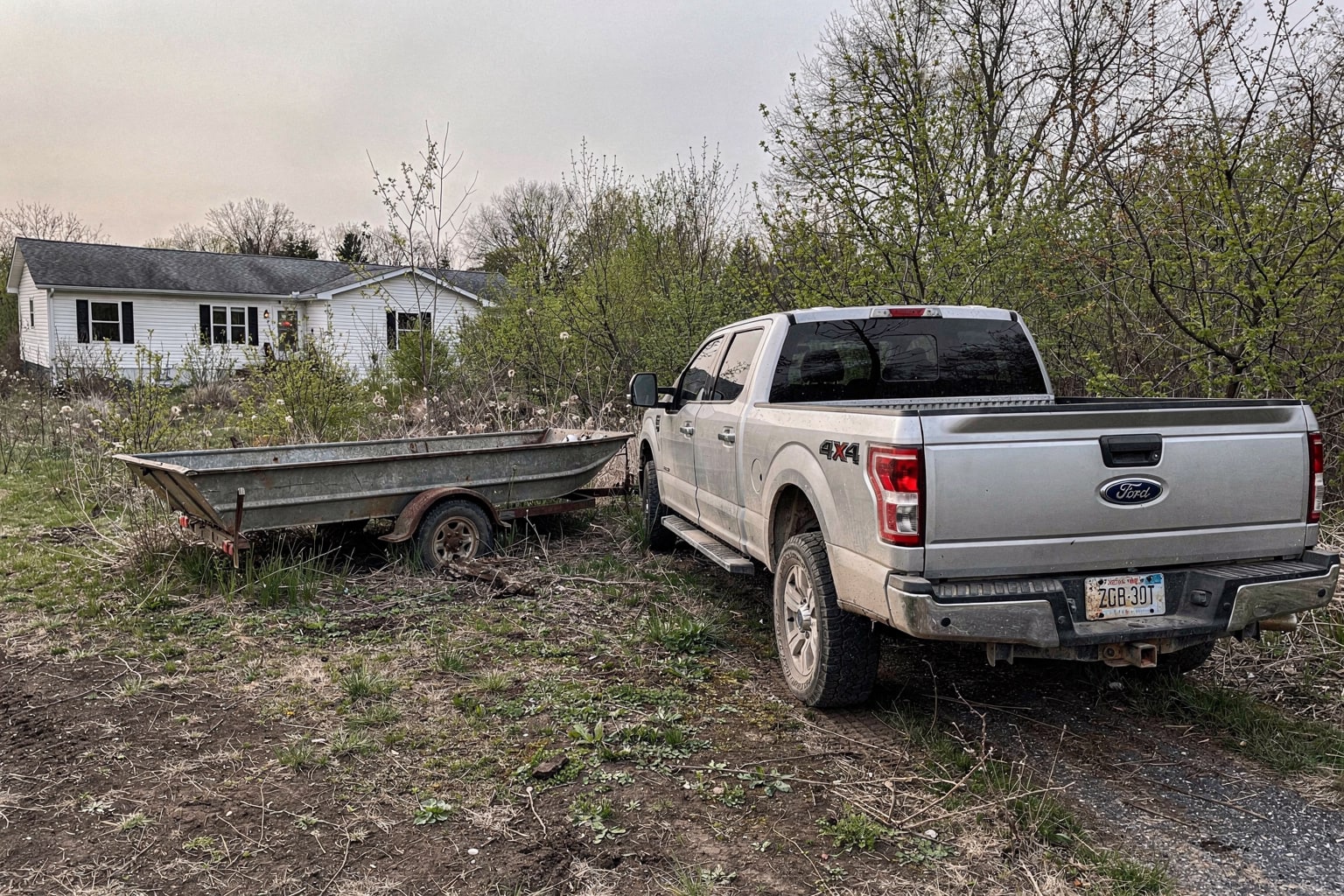 Abandoned weathered aluminum jon boat on overgrown rural lot with kudzu vines, pickup truck and empty trailer approaching for junk boat removal