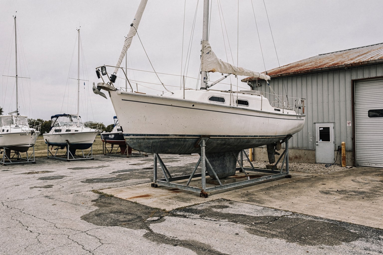 Large 28-foot sailboat on steel cradle in Ohio boatyard with mast removed, other boats and workshop building visible for marine salvage service