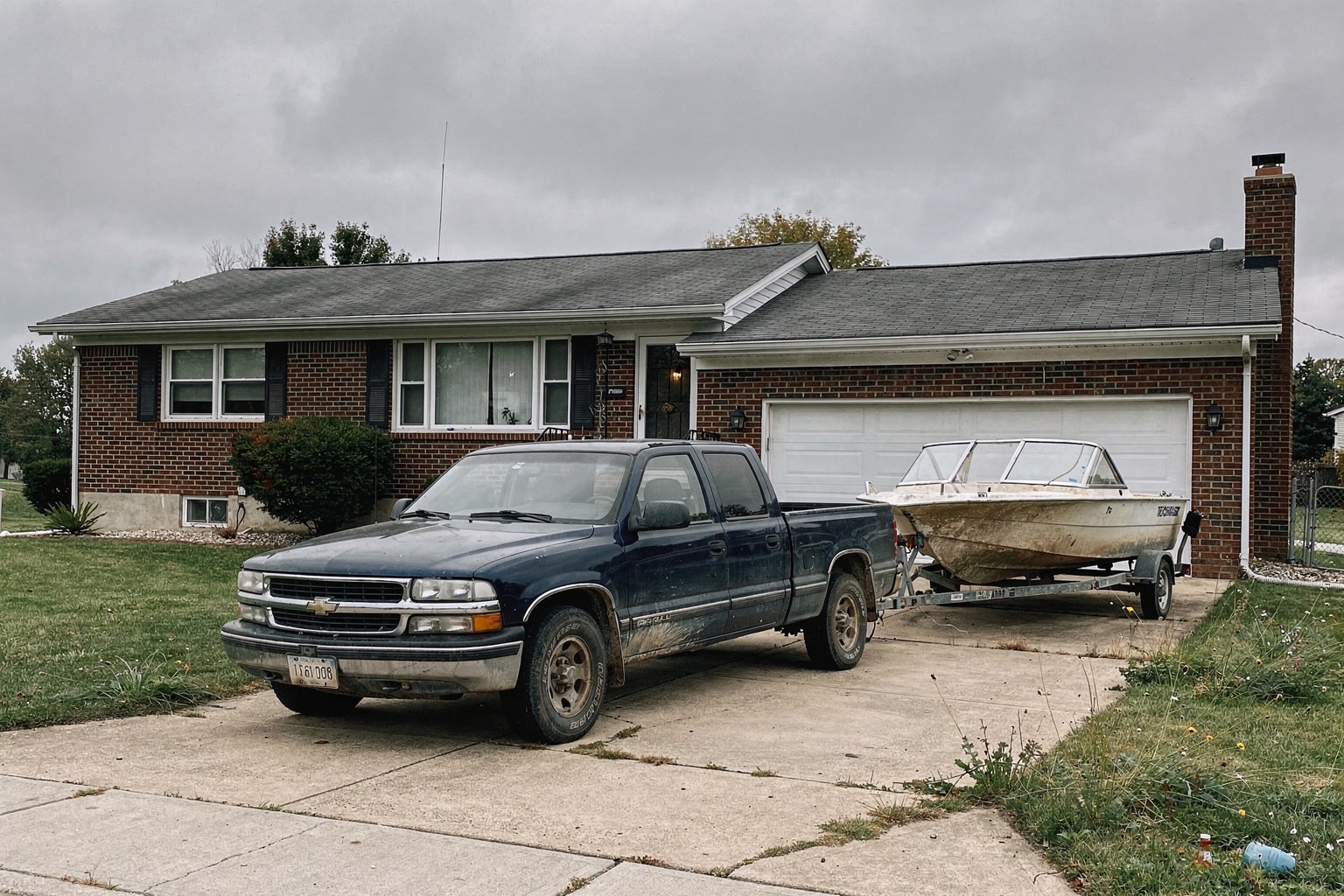 Faded cabin cruiser boat loaded on trailer attached to blue Chevy pickup truck in Ohio residential driveway with brick home and lawn visible
