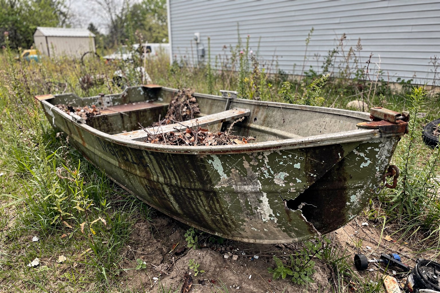 Abandoned junk boat removal needed: weathered aluminum jon boat in overgrown Omaha yard with debris and neglected property