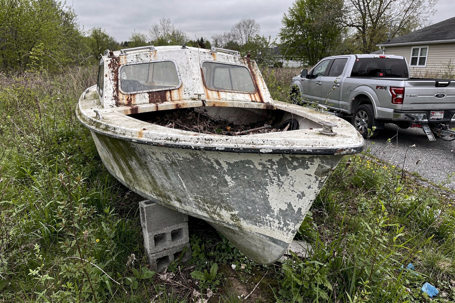 Junk boat disposal Portland Oregon — abandoned fiberglass sailboat on cinder blocks surrounded by overgrown weeds and vines