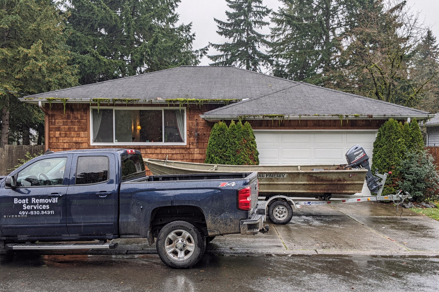 Boat removal Seattle: dark blue pickup truck and trailer carrying weathered aluminum fishing boat on residential driveway