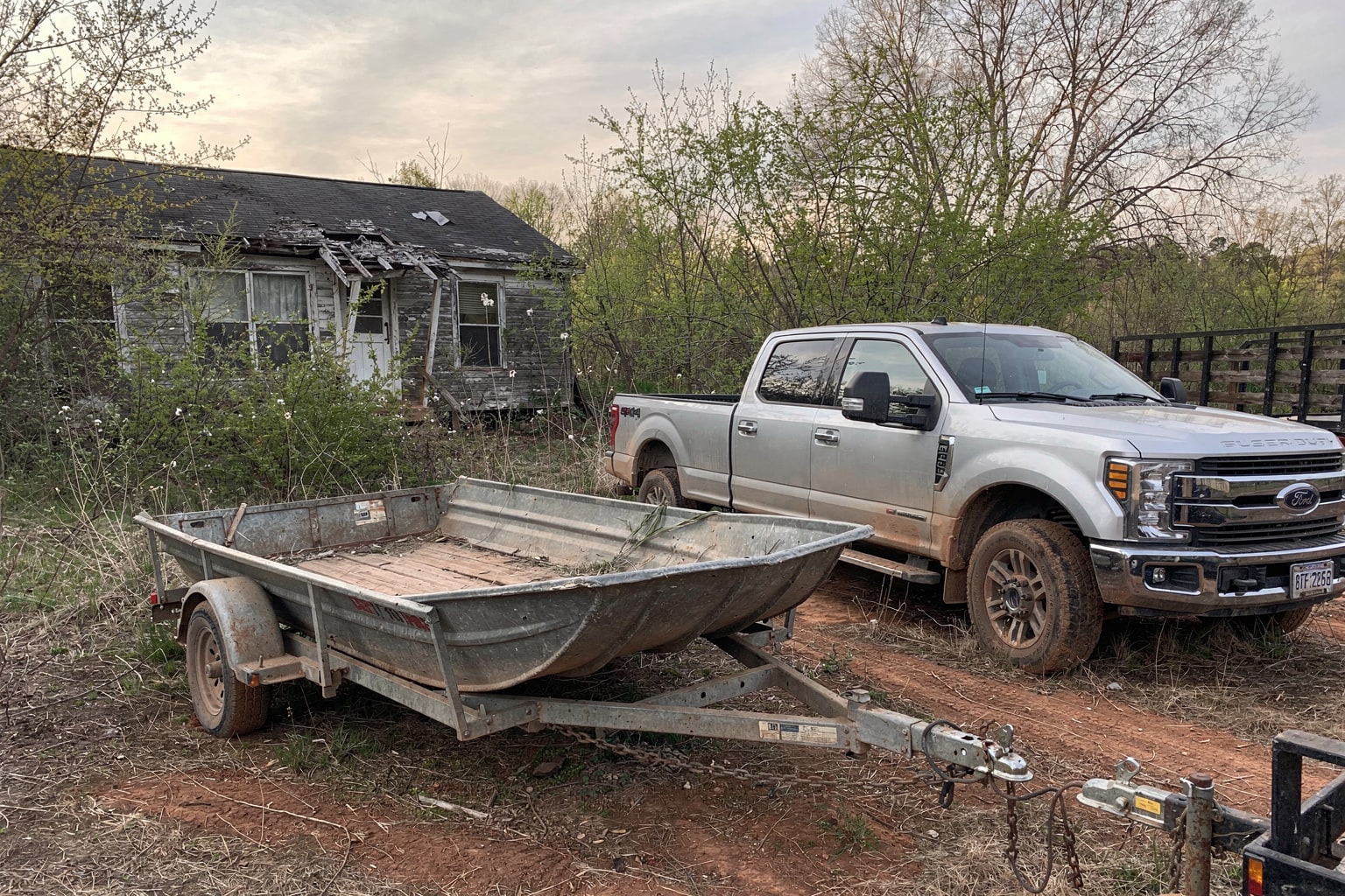 Abandoned aluminum jon boat in overgrown Virginia backyard with pickup truck and trailer approaching for removal