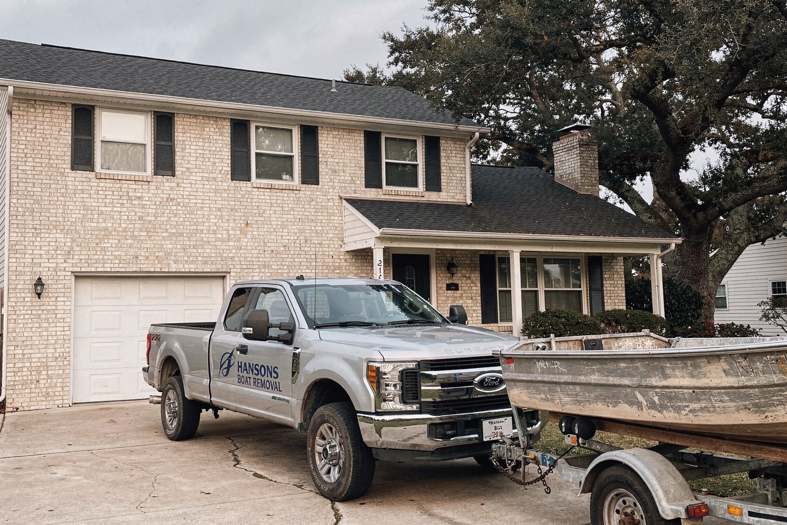 Silver pickup truck with boat removal trailer carrying weathered fishing boat on Virginia Beach residential driveway