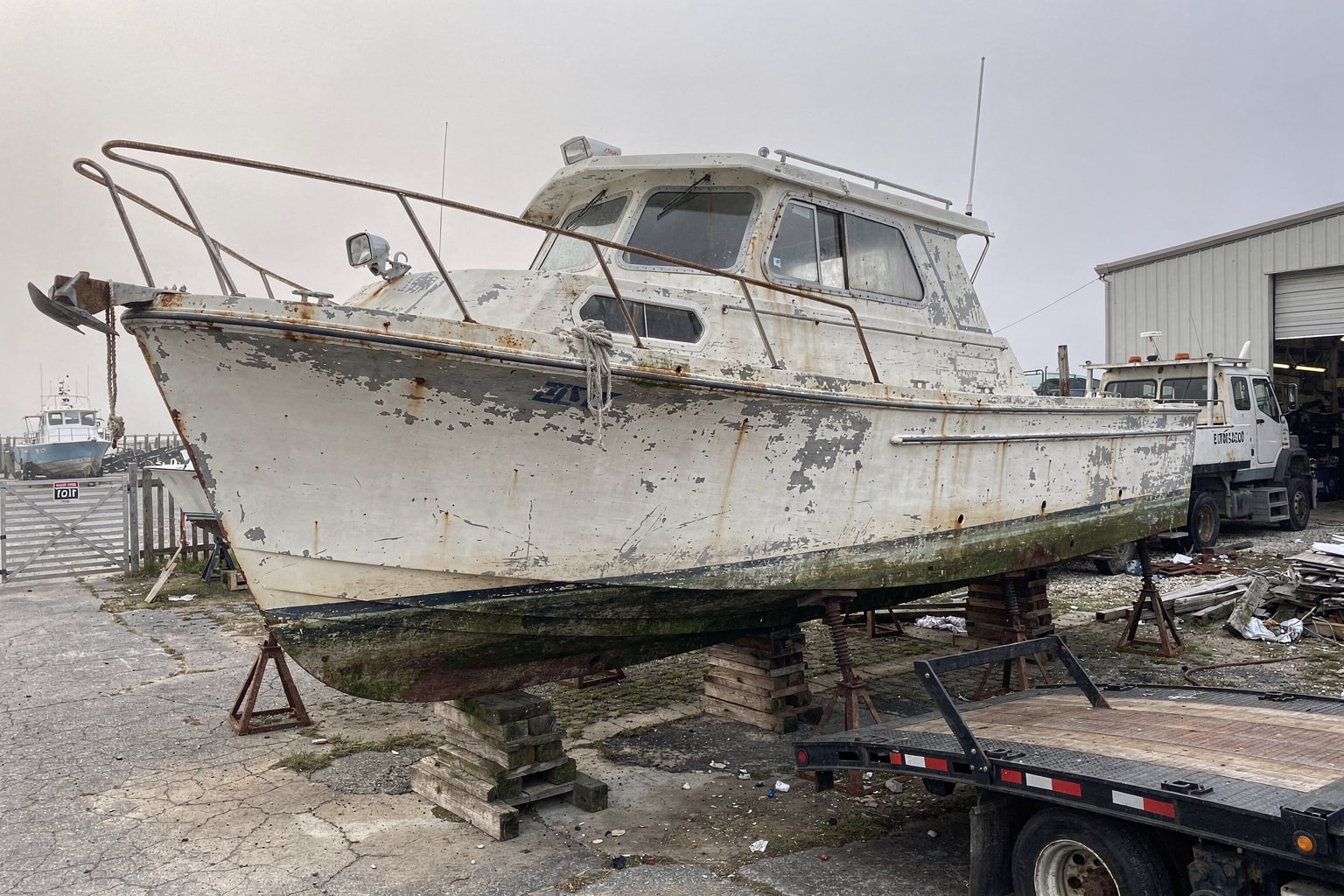 Weathered cabin cruiser boat on blocks in Virginia marine salvage yard with flatbed truck positioned for removal