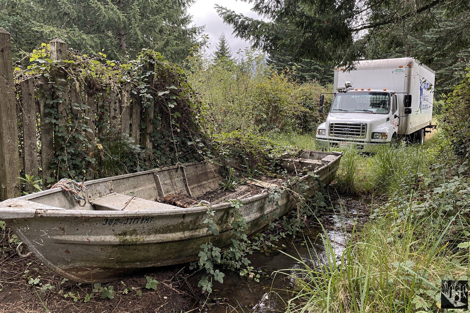 Abandoned jon boat salvage in overgrown Western Washington waterfront yard with old dock pilings and boathouse