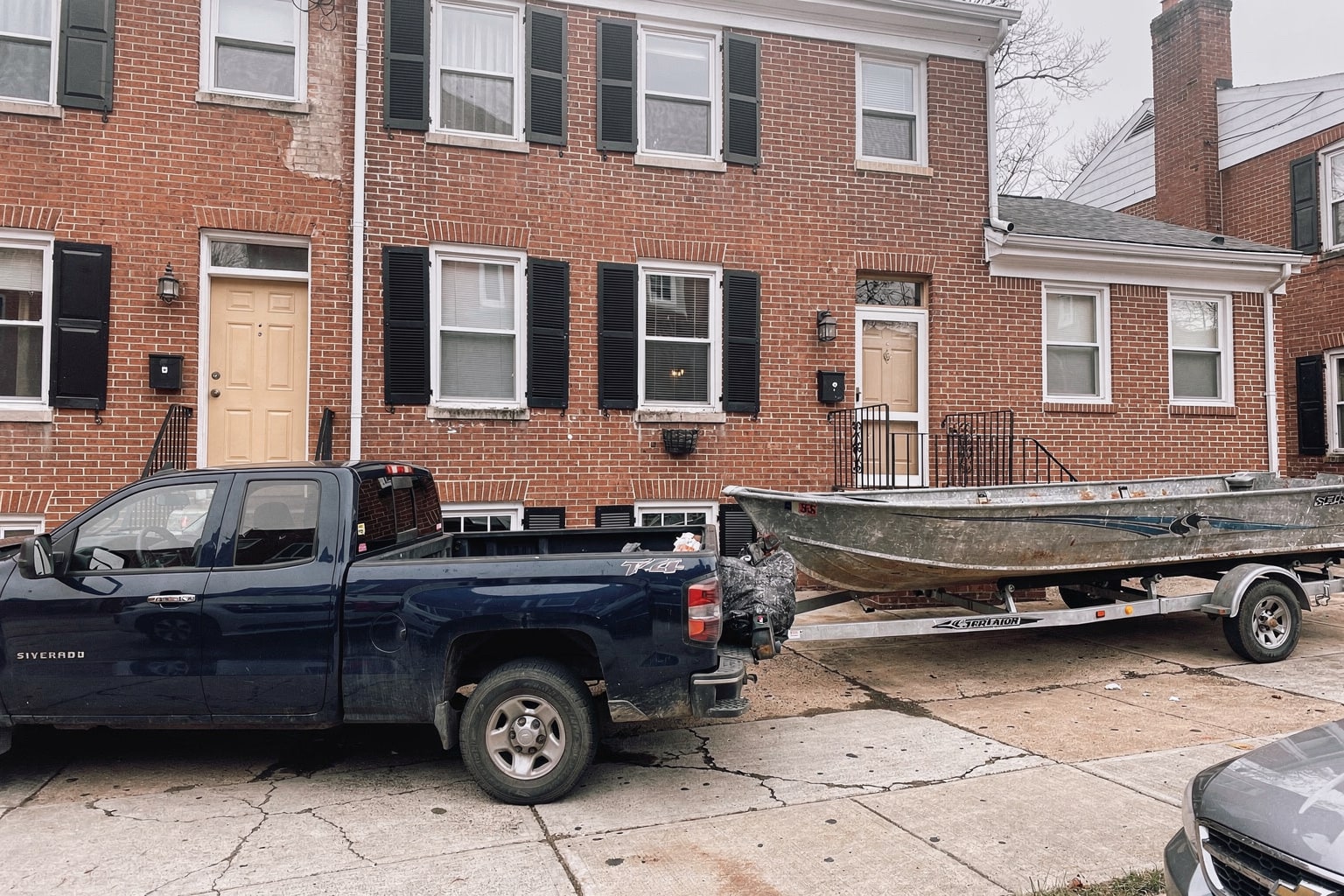 Weathered 16-foot aluminum fishing boat on trailer behind pickup truck, Washington DC residential driveway with brick rowhouse