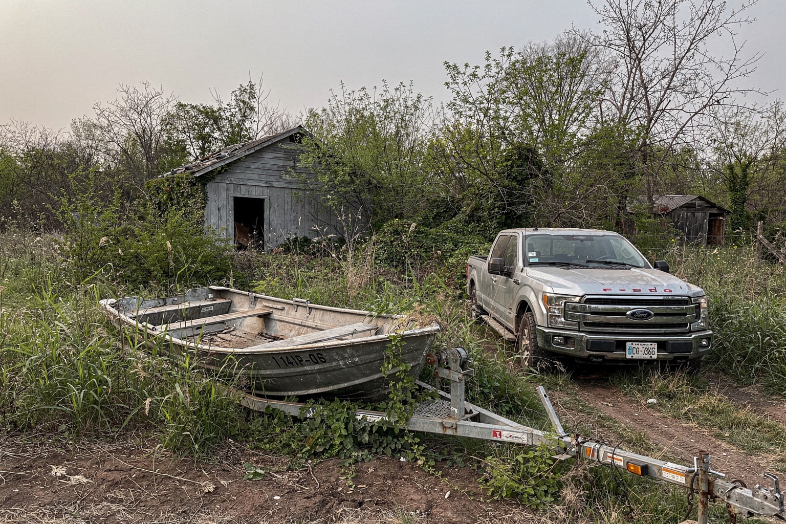 Abandoned 14-foot aluminum fishing boat on rural Wichita property surrounded by overgrown weeds with pickup truck and empty trailer staged for junk boat removal