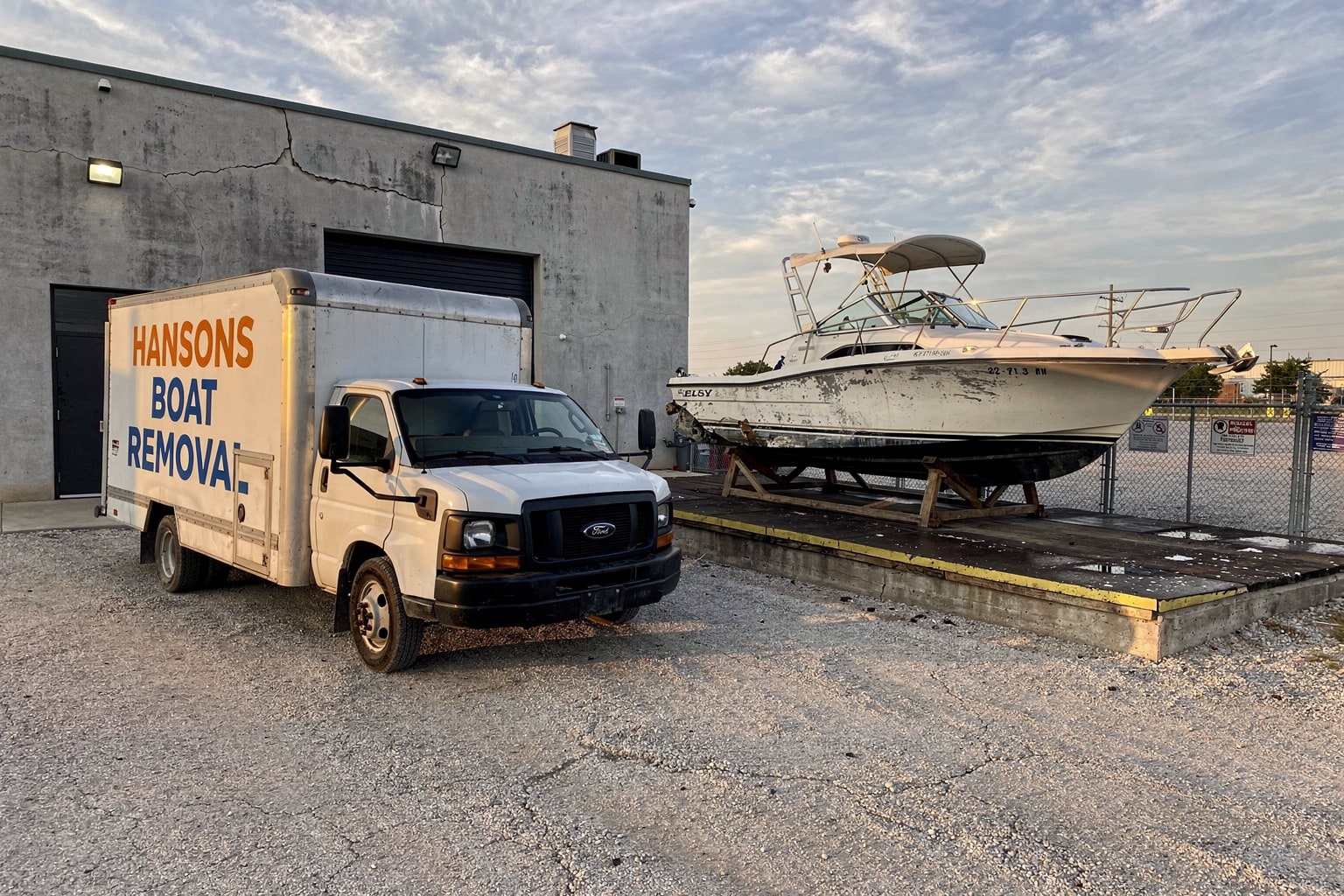 White box truck parked at Wichita commercial lot with weathered 22-foot cabin cruiser sitting on blocks awaiting junk boat removal