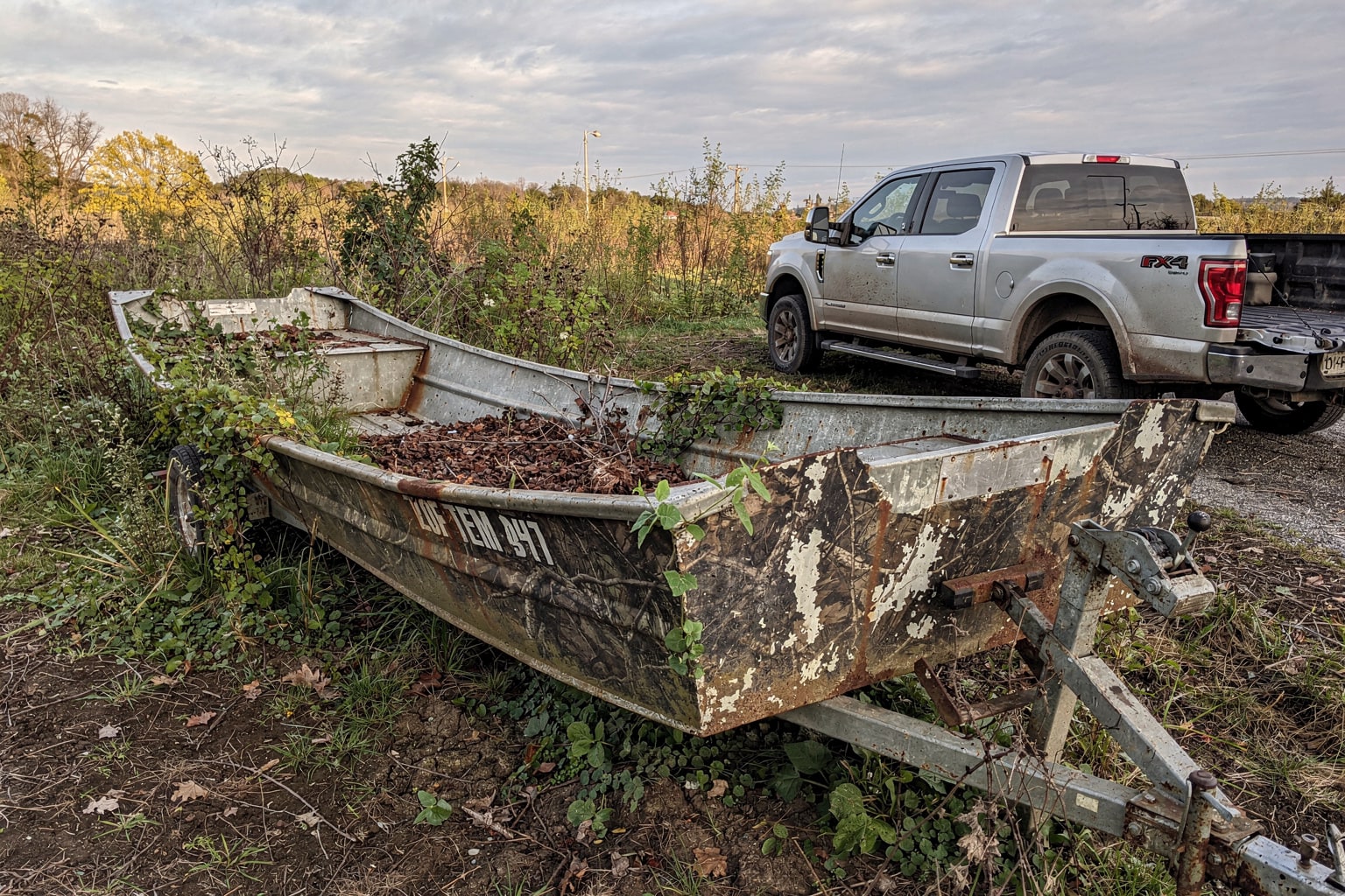 Abandoned weathered jon boat on bare earth in overgrown rural Pennsylvania property with recovery truck