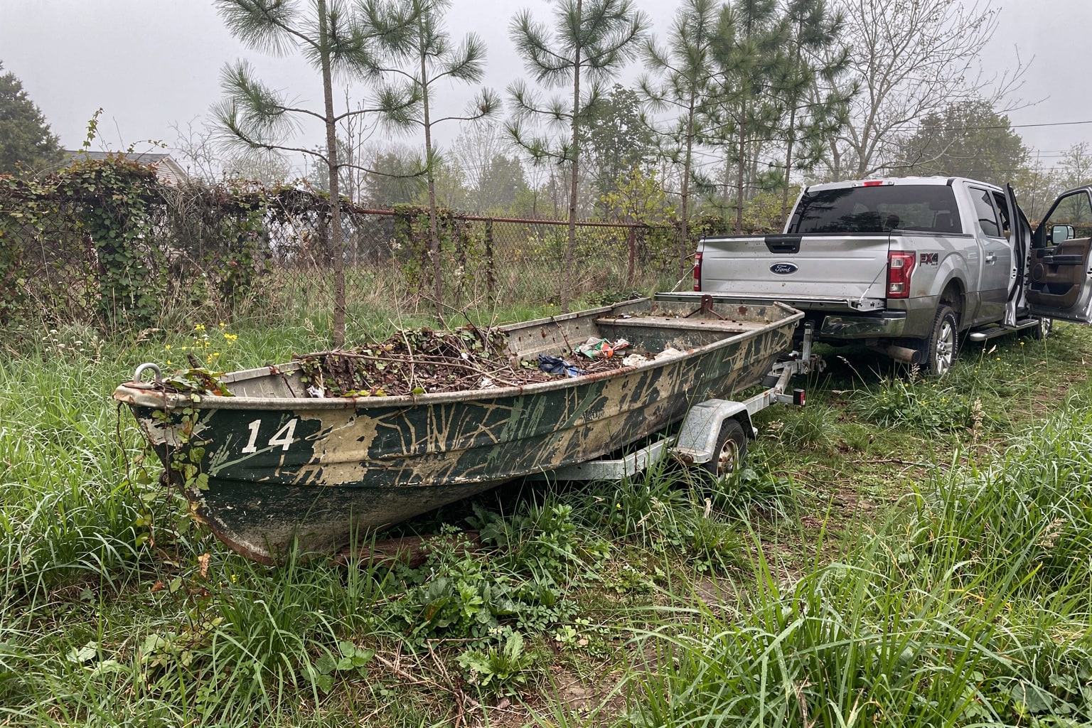 Junk boat removal Brookhaven NY — abandoned 18-foot aluminum fishing boat in overgrown weeds with pickup truck and trailer approaching