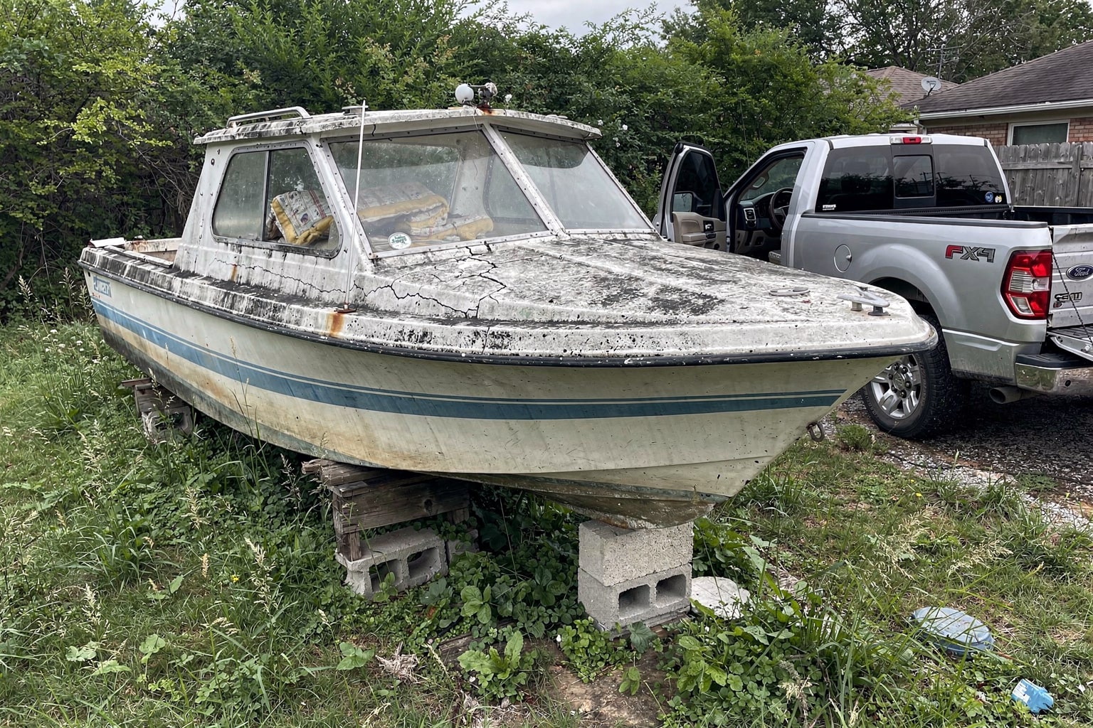 Abandoned weathered cabin cruiser in overgrown Fort Worth backyard surrounded by weeds for junk boat removal