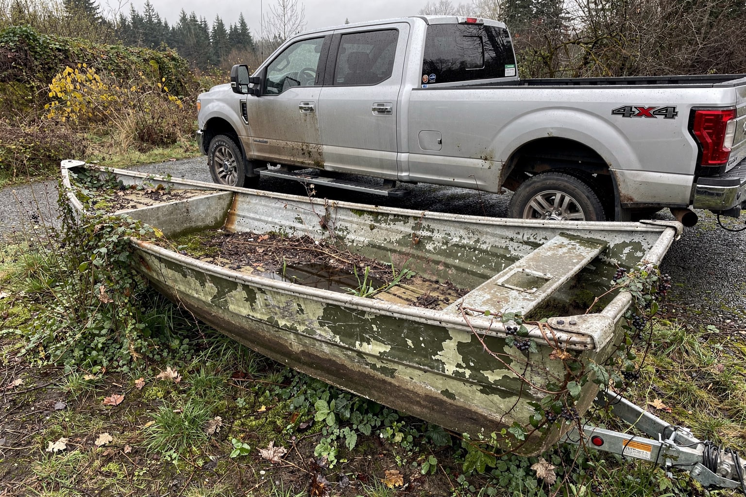 Junk boat removal Seattle: abandoned aluminum jon boat in overgrown backyard with truck and trailer ready for hauling