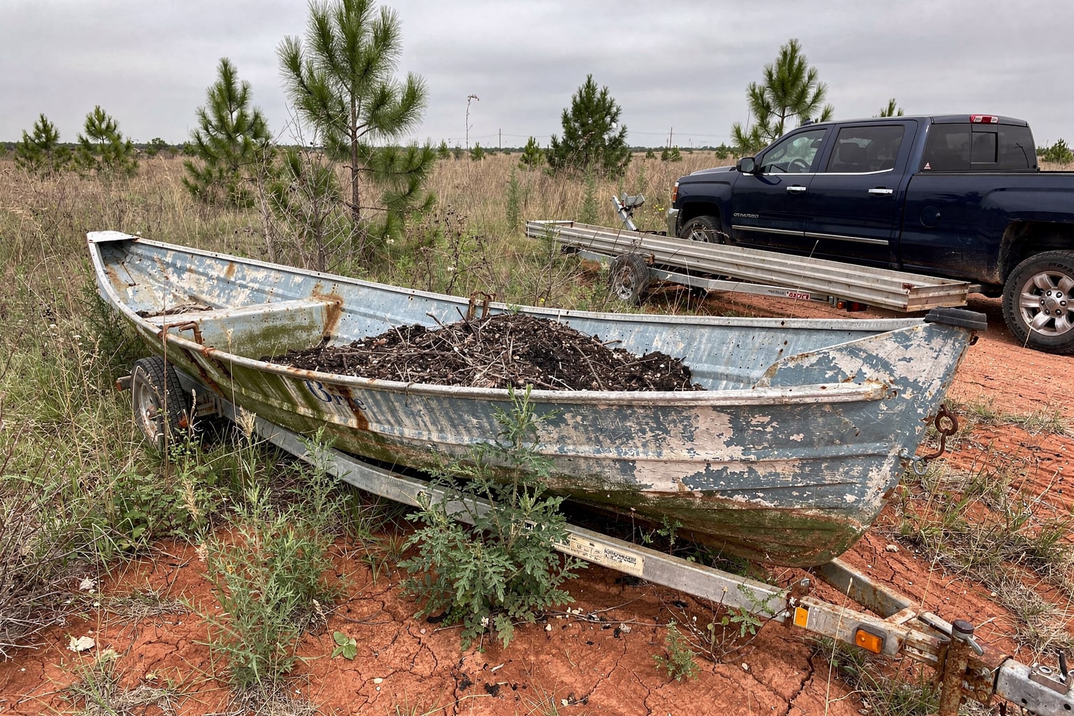Junk boat removal Tulsa Oklahoma - abandoned aluminum fishing boat in overgrown yard with pickup truck and trailer approaching